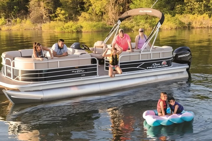 People on a pontoon boat and two kids on a float in a lake with trees in the background.