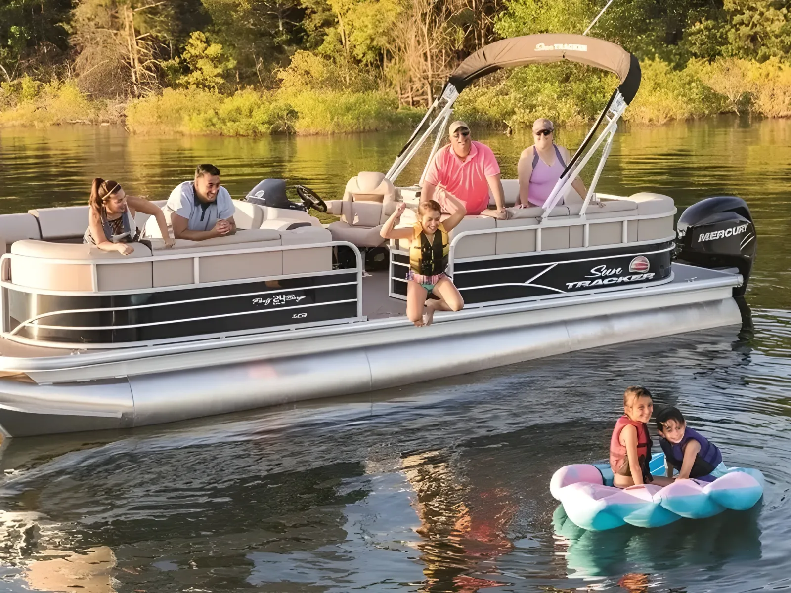 People on a pontoon boat and two kids on a float in a lake with trees in the background.
