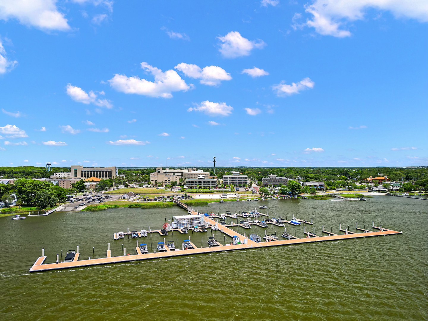 Marina with boats docked on a sunny day, buildings and trees in the background.